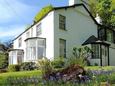 A white house with multiple windows surrounded by green bushes and purple flowers at Loughrigg Cottage in Ambleside