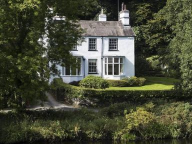A white two-story house with chimneys surrounded by trees and hedges at Loughrigg Cottage in Ambleside