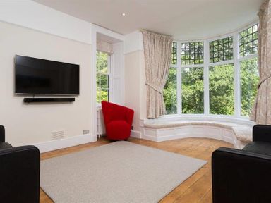 A living room with a wall-mounted television red and black chairs a bay window with curtains and a cushioned seat at Loughrigg Cottage in Ambleside