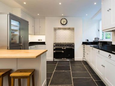 A kitchen with a wooden countertop, white cabinets, black countertops, a black stove under a clock and stone tile flooring at Loughrigg Cottage in Ambleside