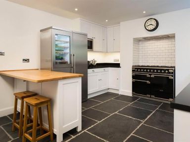 A kitchen with a wooden countertop island and two stools a stainless steel refrigerator white cabinets a black stove set into a tiled alcove and a wall clock at Loughrigg Cottage in Ambleside