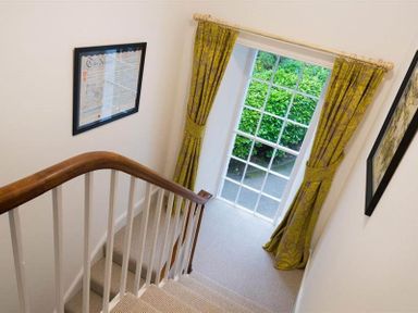 A staircase with wooden handrail and white balusters beside a window with yellow curtains and framed pictures on walls at Loughrigg Cottage in Ambleside