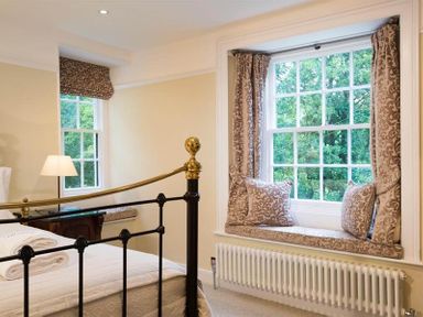 A bedroom with a black and gold metal bed frame a window seat with cushions and curtains and a lamp on a side table at Loughrigg Cottage in Ambleside