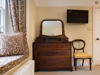 A room with a wooden dresser with mirror a wooden chair with yellow cushion a wall-mounted tv and a window seat with patterned cushions at Loughrigg Cottage Ambleside