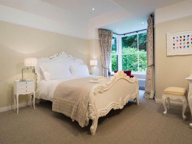 A bedroom with a white ornate bed beige bedding two bedside tables with lamps a window with curtains and a small upholstered stool at Loughrigg Cottage in Ambleside