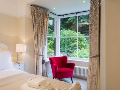 A bedroom with a red armchair next to large windows with patterned curtains at Loughrigg Cottage in Ambleside