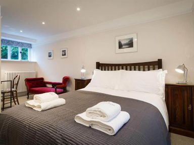 A bedroom with a double bed with white sheets and grey blanket two folded towels two bedside tables with lamps a red chaise lounge and chair by a window at Loughrigg Cottage in Ambleside