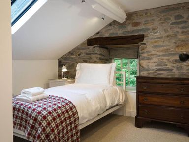 A bedroom with a single bed covered with white bedding and a red patterned blanket a wooden dresser and a stone wall at Loughrigg Cottage in Ambleside