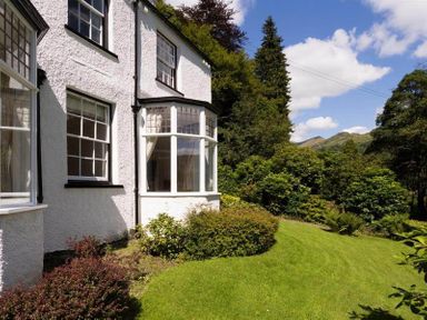 Exterior of a white house with multiple windows surrounded by bushes and a grassy lawn with trees and hills in the background at Loughrigg Cottage in Ambleside