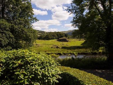 A grassy field with trees and a stream under a partly cloudy sky at Loughrigg Cottage in Ambleside