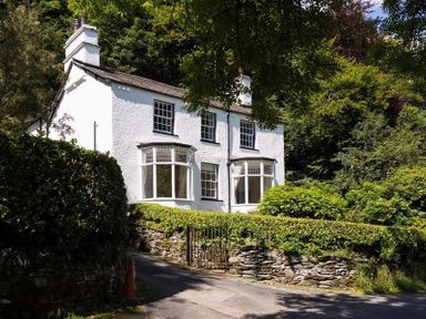 A white two-story house with bay windows surrounded by trees and bushes at Loughrigg Cottage in Ambleside