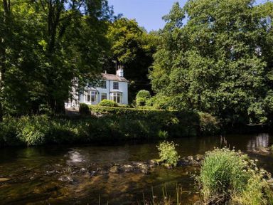 A white house surrounded by trees next to a flowing river at Loughrigg Cottage in Ambleside