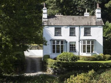 A white two-story house with chimneys surrounded by trees and shrubs at Loughrigg Cottage in Ambleside