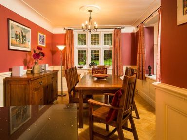 A dining room with wooden table and chairs wooden sideboard floor lamp and paintings on orange walls at Priory Lodge in Windermere