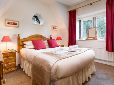 A bedroom with a wooden bed frame two red pillows two bedside tables with lamps and a window with red curtains at Priory Lodge in Windermere