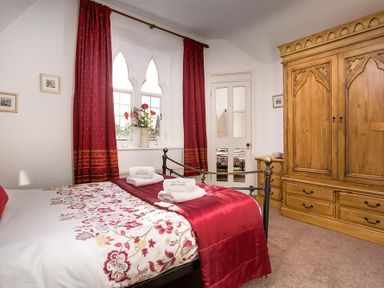 A bedroom with a bed with floral bedding and red pillows a wooden wardrobe and red curtains at Priory Lodge in Windermere