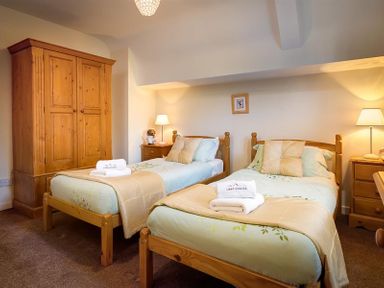 A bedroom with two single beds, wooden wardrobes, bedside tables with lamps, and folded towels at Priory Lodge in Windermere