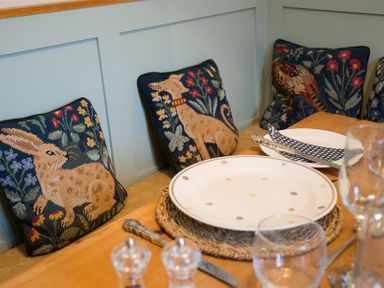 A dining table set with plates glasses and cutlery next to cushions with animal designs on a bench at The Blithe Hare in Troutbeck Bridge