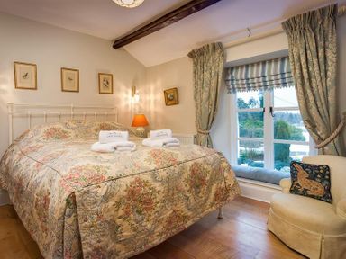 A bedroom with a bed covered in floral bedding beside a window with curtains and a chair with a rabbit cushion at The Blithe Hare in Troutbeck Bridge