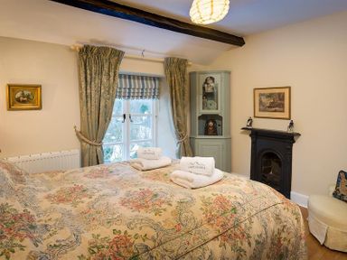 A bedroom with a bed covered with a floral bedspread and folded towels on top a window with curtains a corner cabinet a fireplace and a chair at The Blithe Hare in Troutbeck Bridge