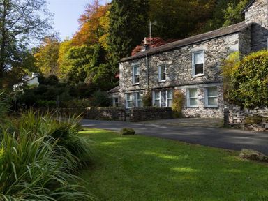 A stone house with multiple windows next to a road and surrounded by trees and grass at The Mews in Ambleside