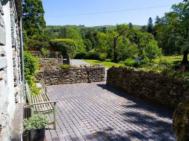 An outdoor stone-paved patio area with a wooden bench and stone walls surrounded by greenery and trees at The Mews in Ambleside