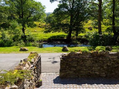 A stone wall with an iron gate opens to a road with trees and green fields behind at The Mews in Ambleside