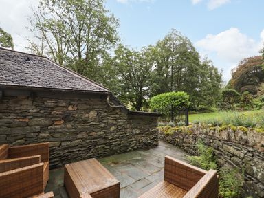 An outdoor stone patio with wicker chairs and a table surrounded by stone walls and trees at The Mews in Ambleside