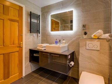 A bathroom with a wooden door a floating vanity with a sink a backlit mirror a towel rack and a toilet at March House in Windermere