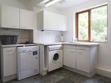 A laundry room with white cabinets a washing machine a dryer a window and a black laundry basket at March House in Windermere