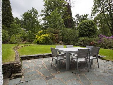 An outdoor patio with a stone floor and a table with six chairs surrounded by a grassy garden with trees and shrubs at March House in Windermere