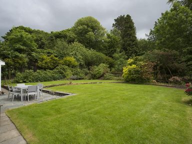 A garden with a lawn and outdoor dining table with chairs surrounded by trees and shrubs at March House in Windermere