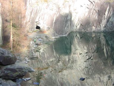 A rocky quarry with a cave entrance and still water reflecting the rock wall at Wythebank in Langdale