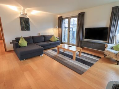 A living room with a dark sofa green cushions wooden coffee table a striped rug a wall-mounted television and large windows with curtains at Idle Mill 1 Ambleside