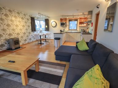 An open plan living area with a dark sofa and green cushions a wooden coffee table a dining table with white chairs and a kitchen with appliances at Idle Mill 1 in Ambleside