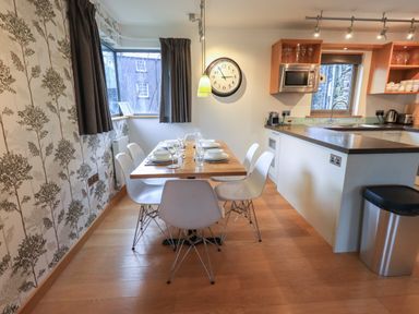 A kitchen and dining area with a wooden floor white chairs a wooden table set with dishes and glasses a wall clock and kitchen appliances at Idle Mill 1 Ambleside