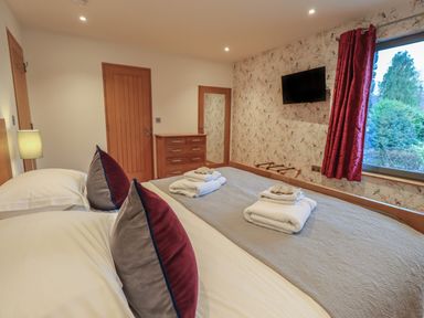 A bedroom with two beds with pillows and towels a chest of drawers a wall mounted television and a window with red curtains at Idle Mill 1 in Ambleside