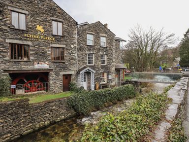 A stone building housing The Flying Fleece pub next to a flowing river with a bridge in the background at Idle Mill 1 in Ambleside