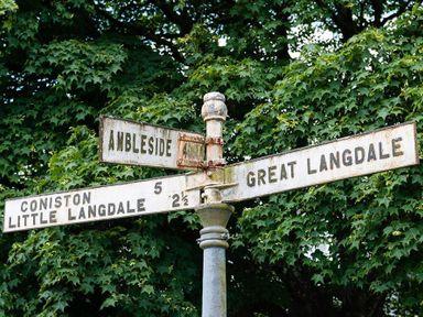 A vintage metal signpost with directions to Ambleside, Great Langdale, Coniston, and Little Langdale with green leafy trees in the background at Le Chalet in Elterwater
