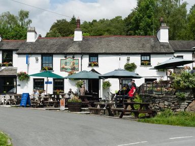 Outdoor seating area with tables umbrellas and people in front of a white building with flower baskets at Le Chalet in Elterwater