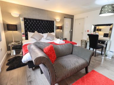 A bedroom with a striped loveseat red cushions a bed with a tufted headboard and mirrored side tables at Le Chalet in Elterwater