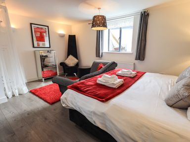 A bedroom with a bed with two sets of towels on a red runner a black chair with a white cushion a mirrored chest of drawers and a window with dark curtains at Le Chalet in Elterwater