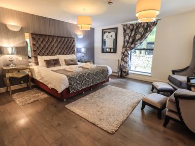 A bedroom with a large bed with a tufted headboard two bedside tables lamps a pair of armchairs ottomans a window with patterned curtains and two rugs on wooden floor at Le Chalet in Elterwater