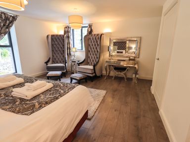 A bedroom with a bed, two high-backed chairs with footstools, a mirrored dressing table with a lamp, and windows with curtains at Le Chalet in Elterwater