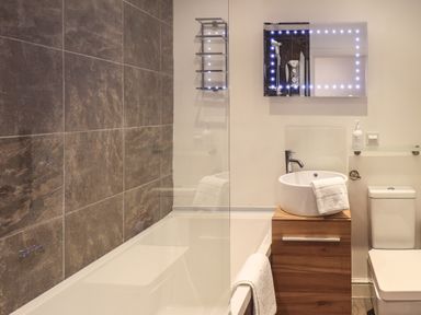 A bathroom with a bathtub tiled wall sink on a wooden cabinet illuminated mirror and a toilet at Le Chalet in Elterwater