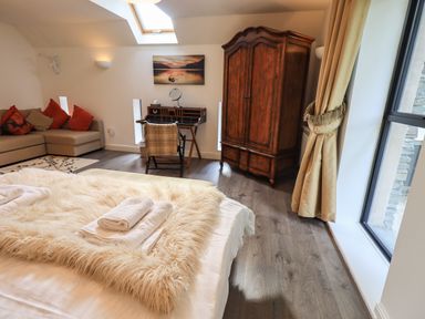 A bedroom with a bed covered with a fur throw and towels a wooden wardrobe a desk with a chair a sofa with cushions and a window with gold curtains at Le Chalet in Elterwater