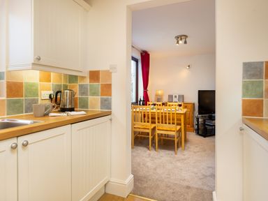 A kitchen with a kettle and mug on the counter leading to a dining area with a wooden table and chairs and a television at High Lake in Bowness