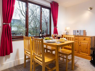 A dining area with a wooden table and chairs next to a window with red curtains at High Lake in Bowness
