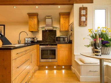 A kitchen with wooden cabinets toaster microwave oven and a small dining table with flowers and a bottle of champagne at Ivy Cottage in Ambleside