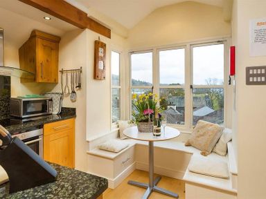A kitchen corner with wooden cabinets a microwave utensils hanging on the wall and a small dining nook with cushioned bench seating and a round table with flowers at Ivy Cottage in Ambleside